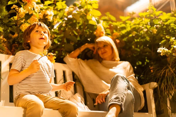 Chłopiec i jego mama siedzą na ławce w parku miejskim. Syn i matka bawią się na lodzie z krzakami w tle. Rodzinny wypoczynek w Autumn Park. Weekend jesienny — Zdjęcie stockowe