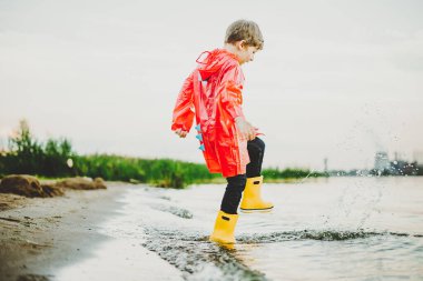 Kırmızı yağmurluklu ve sarı lastik çizmeli bir çocuk sahilde suyla oynuyor. Su geçirmez montlu okul çocuğu denizde suya atlıyor. Çocuk sahilde dalgalarla eğleniyor.