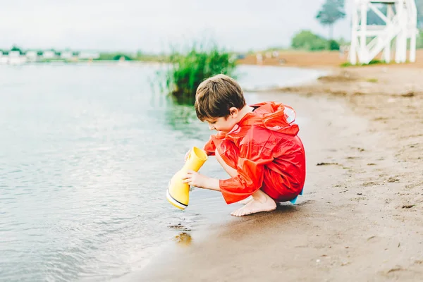 Chłopiec w czerwonym płaszczu wlewa wodę z żółtego gumowego buta do jeziora. Dziecko bawiące się wodą w stawie. Dzieciak bawiący się na świeżym powietrzu w pobliżu rzeki — Zdjęcie stockowe