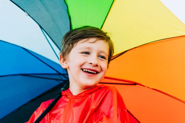 Portrét rozesmátého školáka s duhovým deštníkem za zády. Usměvavé dítě drží na rameni barevný deštník. Veselé dítě v červeném plášti, držící vícebarevný deštník — Stock fotografie