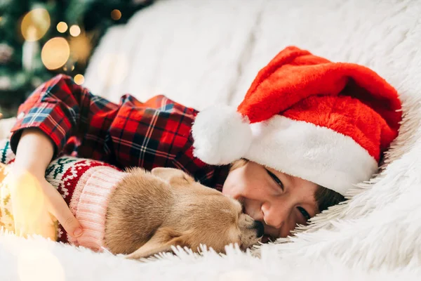 Feliz Navidad. Niño y perrito feliz en suéter jugando, abrazándose y divirtiéndose en el sofá con manta en las vacaciones de invierno. Niño con sombrero de Santa Claus. Árbol de Navidad con luz bokeh. Feliz año nuevo — Foto de Stock