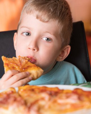 Güzel, mutlu, taze pizza dilimini ısıran genç bir çocuk siyah koltukta oturuyor, sarı saçları var..