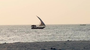 İki yelkenli (Dhows) gün batımında dalgalar arasında yarışıyor, sudan yansıyan altın güneş ışığı okyanusun dingin ve canlı bir atmosferini yaratıyor..