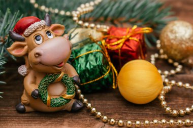 Noel Baba şapkalı oyuncak boğa koyu tahta bir arka planda. Arka planda küçük hediye kutuları ve topları olan Noel ağacı dalı. 2021 yılının sembolü olarak boğa, Noel konsepti..