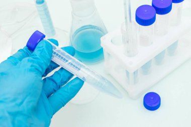 Mavi bir tıbbi eldiven, üzerinde mavi sıvı bulunan bir test tüpü taşır. Arka planda mavi sıvı bulunan bir tripod. Kimyasal teknoloji kavramı, biyoteknoloji, eczacılık.