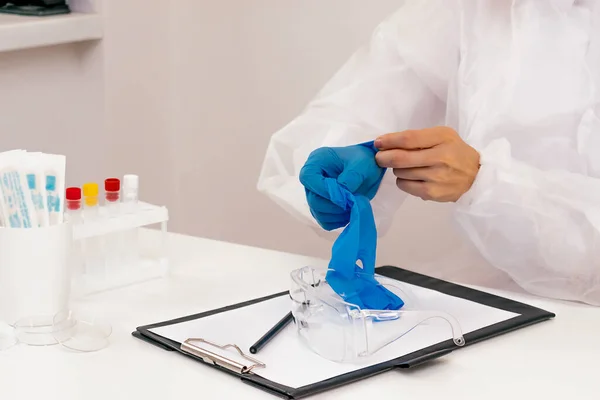 Doctor in a protective suit in the medical office takes off his latex gloves.Safety glasses and writing paper on the table.Coronavirus concept.