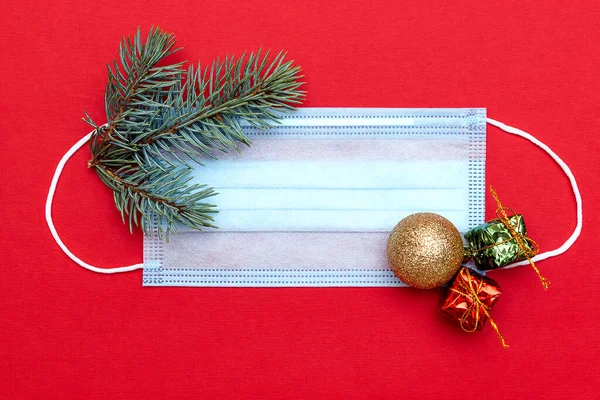 Medical protective mask on a red background.The mask is decorated with a fir branches,ball and little gift boxes.New Year,Christmas and coronavirus concept.Copy space