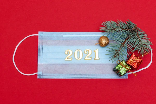 Medical protective mask on a red background.The mask is decorated with a spruce branch, small gift boxes and the numbers 2021.New Year and coronavirus concept.