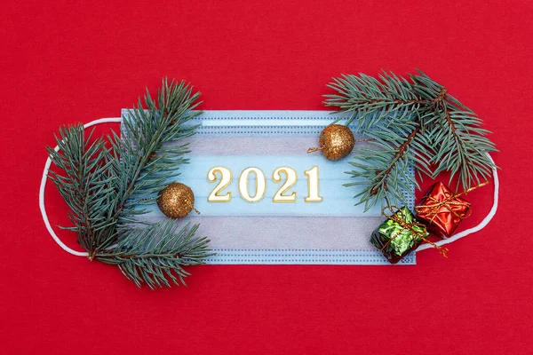 Medical protective mask on a red background.The mask is decorated with a spruce branch, small gift boxes and the numbers 2021.New Year and coronavirus concept.