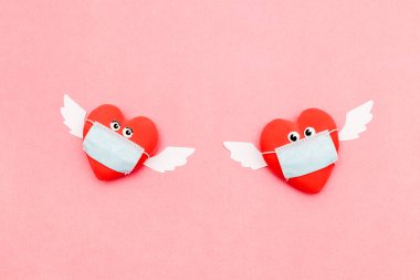 Pembe arka planda gözleri ve melek kanatları olan koruyucu maskeli iki kalp. Sevgililer Günü ve koronavirüs salgını. Düz konsept, üst görünüm..