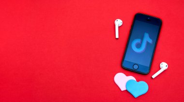 Üzerinde Tik Tok logosu olan akıllı bir telefon, kablosuz kulaklıklar ve kırmızı arka planda iki renkli kalp. Sevgililer Günü ve teknoloji konsepti. Boşluğu, manzarayı, düzlüğü, düzlüğü kopyala..