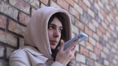 Gülümseyen esmer bir kız telefonuna sesli mesaj kaydediyor. Arka planda kahverengi tuğla bir duvar. Düşük hareket..