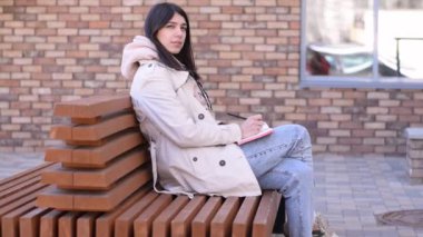 Pozitif esmer kız not defterine not alıyor. Sokakta bir bankta. Düşük hareket..