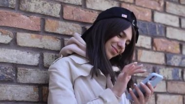 Siyah şapkalı, gülümseyen esmer bir kız telefonunda bir sesli mesaj kaydediyor. Arka planda kahverengi tuğla bir duvar..