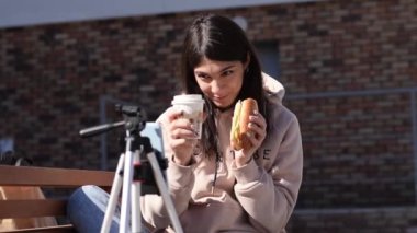 Esmer kız hamburger yiyor ve sosyal ağlardan canlı yayın yapıyor. Akıllı telefon tripoda takılı. Takipçilerle iletişim, serbest çalışma, online toplantı