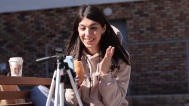 Esmer kız hamburger yiyor ve sosyal ağlardan canlı yayın yapıyor. Akıllı telefon tripoda takılı. Takipçilerle iletişim, serbest çalışma, online toplantı