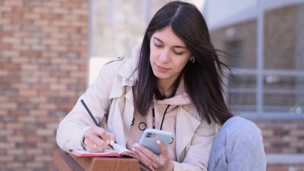 Ευτυχισμένη Μελαχρινή Κοπέλα Χρησιμοποιώντας Ένα Smartphone Και Κρατώντας Σημειώσεις Ένα — Αρχείο Βίντεο