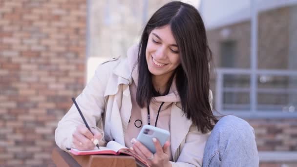 Menina Morena Feliz Usando Smartphone Fazendo Anotações Caderno Banco Rua — Vídeo de Stock