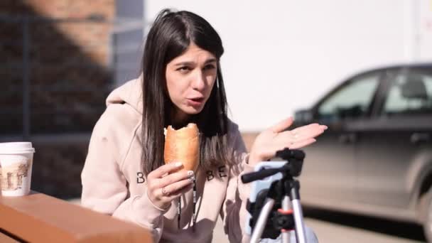 ブルネットの女の子はハンバーガーを食べて 路上のベンチでソーシャルネットワーク上のライブ放送を記録します スマートフォンは三脚に取り付けられています フォロワーとのコミュニケーション フリーランス オンライン会議 — ストック動画