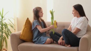 Kız, anneler gününde annesini tebrik eder ve ona çiçekler ve ev yapımı bir hediye verir. Evdeki kanepede otururken sarılıp gülüyorlar. Anneler Günü, 8 Mart ve Doğum Günü konsepti.