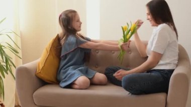 Kız, anneler gününde annesini tebrik eder ve ona çiçekler ve ev yapımı bir hediye verir. Evdeki kanepede otururken sarılıp gülüyorlar. Anneler Günü, 8 Mart ve Doğum Günü konsepti.