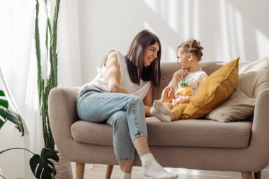 A dark-haired young mixed-race woman plays on the couch with her little daughter.Home interior design with houseplants.Time together.Family concept and Mother's Day concept. clipart
