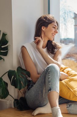 Koyu saçlı, melez bir kız pencerenin kenarında oturuyor ve güneşli günün tadını çıkarıyor. Ev tasarımları. Pencerenin dışındaki şehir manzarası. Rahatlama ve iyi ruh hali kavramı..