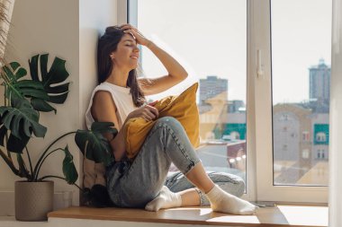 Koyu saçlı, melez bir kız pencerenin kenarında oturuyor ve güneşli günün tadını çıkarıyor. Ev tasarımları. Pencerenin dışındaki şehir manzarası. Rahatlama ve iyi ruh hali kavramı..