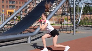 Tişörtlü, şortlu ve spor ayakkabılı atlama hareketleriyle fonksiyonel bir egzersiz yapan atletik kız bacaklarını ve dambıllarını sallıyor. Sağlıklı yaşam tarzı. Açık hava eğitimi..