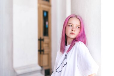 Beyaz tişörtlü pembe saçlı genç bir kızın portresi bir binanın beyaz duvarına dayanıyor. Sokak stili. Z nesli tarzı. Metin için alan kopyala.