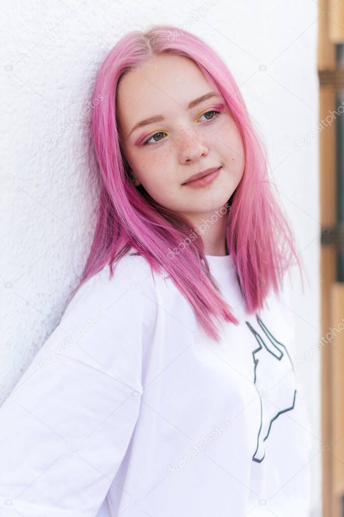 Portrait of a positive pink-haired teenage girl in a white T-shirt leaning against the white wall of a building.Street style.Summer concept.Generation Z style.