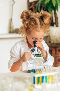 Beyaz üniformalı ve koruyucu gözlüklü küçük bir kız laboratuardaki mikroskopa bakar. Okula geri döner. Genç bilim adamları. Anaokulu ve çocukların okul eğitimi..