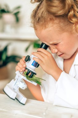 Beyaz üniformalı ve koruyucu gözlüklü küçük bir kız laboratuardaki mikroskopa bakar. Okula geri döner. Genç bilim adamları. Anaokulu ve çocukların okul eğitimi..