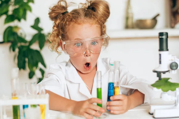 Surprised little red-haired girl in white uniform conducting chemical experiments in a laboratory.Back to school concept.Young scientists.Natural sciences.Preschool and school education of children.