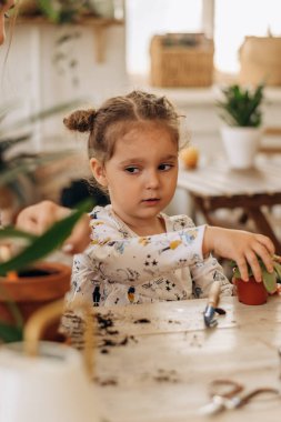 Annesiyle birlikte küçük melez, koyu renk saçlı kız evdeki teras saksılarına çiçek dikiyor. Aile eğlencesi, bahçe işleri, hobi anlayışı. Biyofili tasarımı ve kentsel orman konsepti..