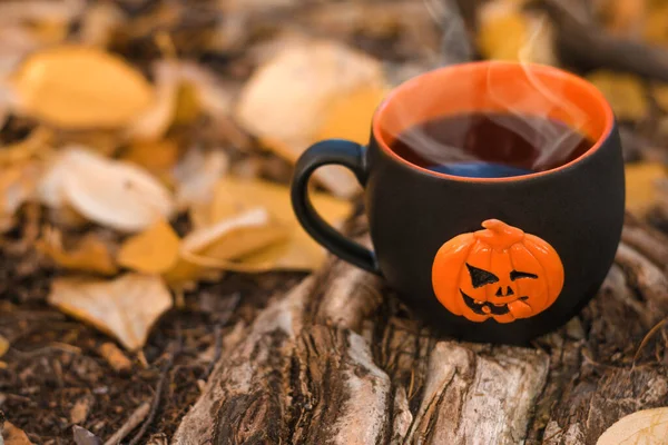 Sıcak çaylı siyah kupa, düşmüş sarı sonbahar yaprakları ve dalları arasında yerde duruyor. Kupa, balkabağı Jack O 'Lantern ile süslenmiş. Cadılar Bayramı kavramı..