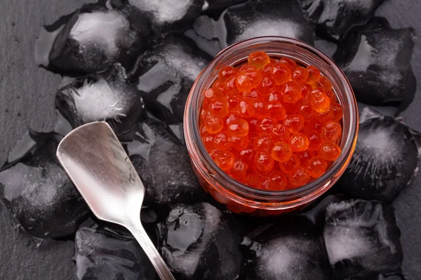 Glass jar with salmon caviar standing in the middle of ice cubes on black slate plate — Stock Photo, Image