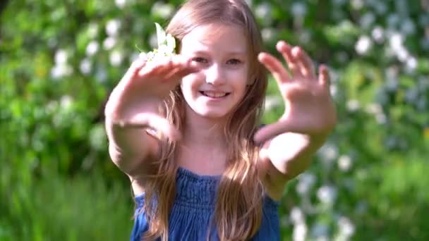Una chica con una flor en pelo largo y rubio muestra un gesto de corazón con las manos en un día soleado al aire libre. Enfoque selectivo — Vídeo de stock