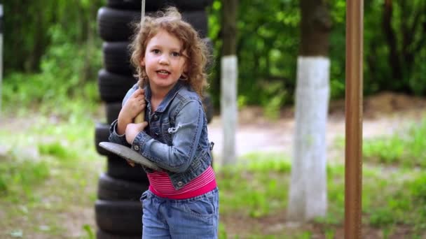 Pequeña chica rizada de 4 años en un traje de mezclilla monta un columpio en verano — Vídeo de stock