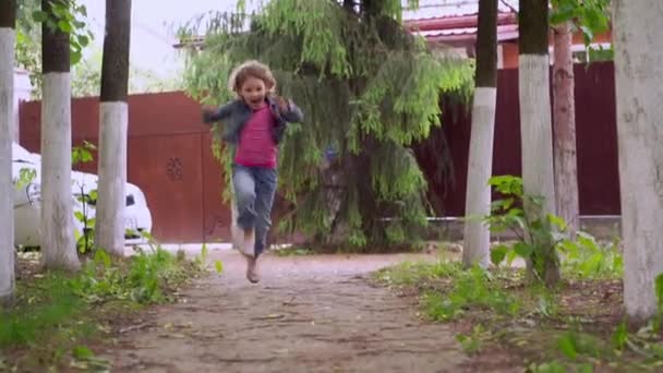 Een klein krullend blond gelukkig meisje van 4-5 jaar rennen en springen langs een pad. — Stockvideo
