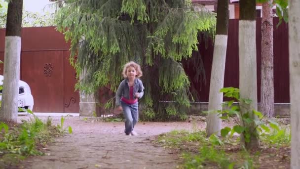Een klein krullend blond gelukkig meisje van 4-5 jaar rennen en springen langs een pad. — Stockvideo