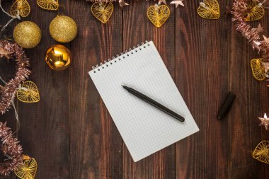 Tahta bir masanın üzerinde noel topları ve çelenk olan not defteri ve siyah kalem. Düz Yatay