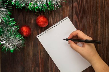 El, yeni yıl topları ve çelenklerle dolu tahta bir masanın üzerinde duran bir defterde siyah bir kalemle yazıyor. Düz Yatay