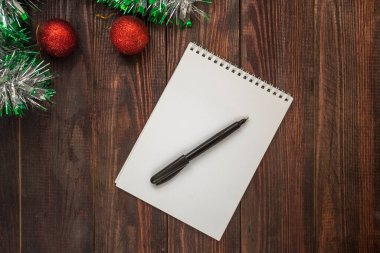 Tahta bir masanın üzerinde noel topları ve çelenk olan not defteri ve siyah kalem. Düz Yatay