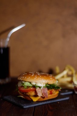 Dikey çerçeve. Fast food. Tavuk pirzolası, domates, peynir, yeşillik ve pastırma. Susam tohumlu çörek. Arka planda patates kızartması ve kola..