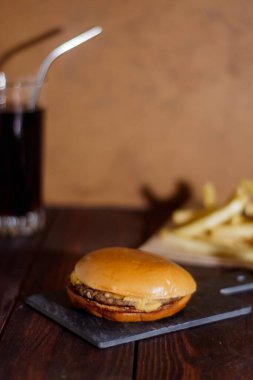 Dikey çerçeve. Fast food. Tavuk pirzolası, domates, peynir, yeşillik. Susam tohumlu çörek. Arka planda patates kızartması ve kola..