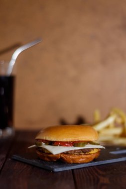 Dikey çerçeve. Fast food. Tavuk pirzolası, domates, peynir, yeşillik. Susam tohumlu çörek. Arka planda patates kızartması ve kola..