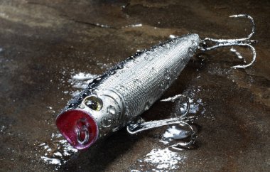 Yapay gümüş ve kırmızı balık yemi. Islak ve ıslanmış bir kayanın üzerinde duran suyun tepesine yerleştirilmiş üç kancalı yem..