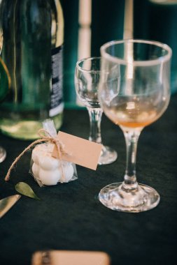 Küçük beyaz, baloncuk şeklinde plastik kaplı, sicim fiyonklu ve boş etiketli, şarap bardaklarının yanındaki masa örtüsünün üzerine yerleştirilmiş.