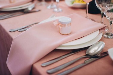 Pembe masa örtüsü ve peçeteyle nefis bir yemek masası seti. Dekoratif konuk ikramiyesi kavanozu, kurdeleyle bağlanmış.